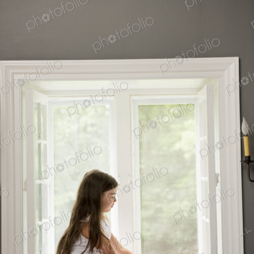 A girl in a white dress seated by a window playing.