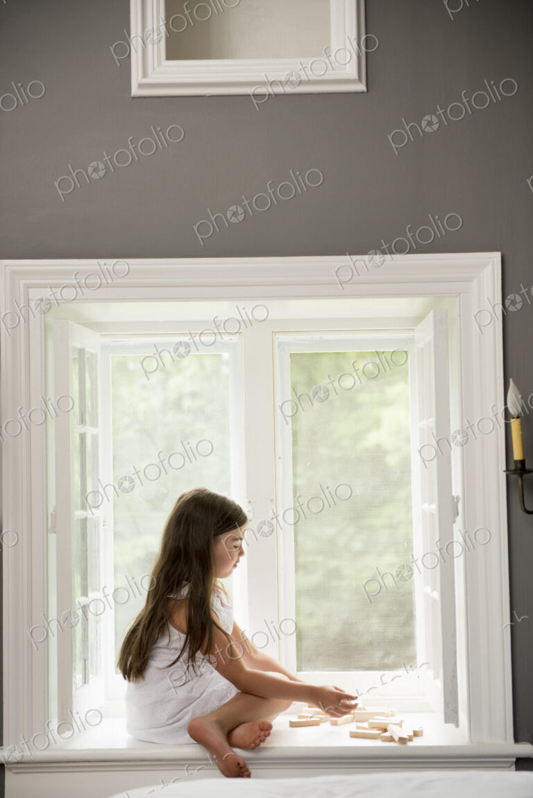 A girl in a white dress seated by a window playing.