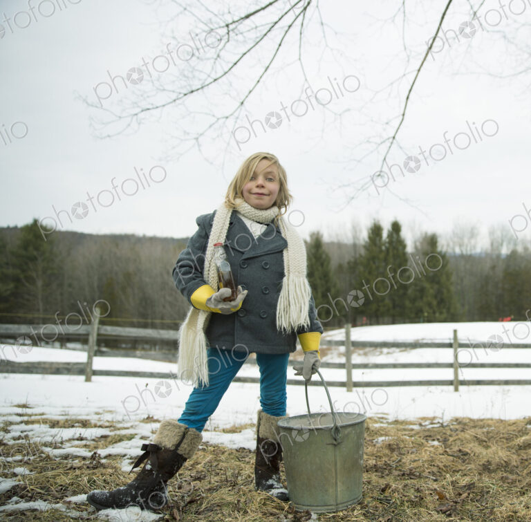 A girl in the snow, carrying a metal bucket.