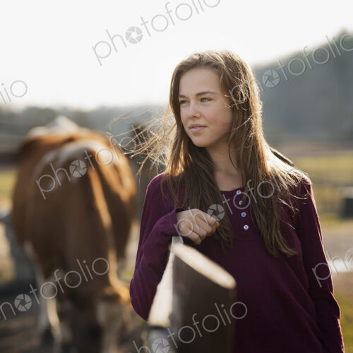 A girl leaning on a paddock fence, and a grazing horse in the background, on an… A girl leaning on a paddock fence, and a grazing horse in the background, on an…