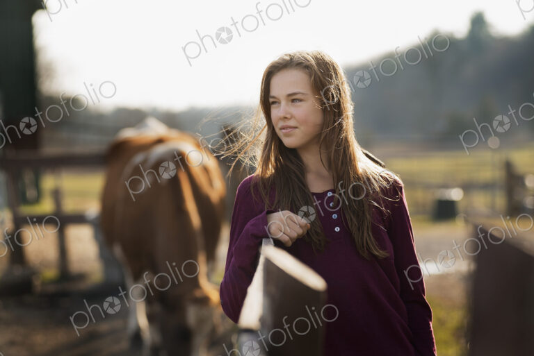 A girl leaning on a paddock fence, and a grazing horse in the background, on an…