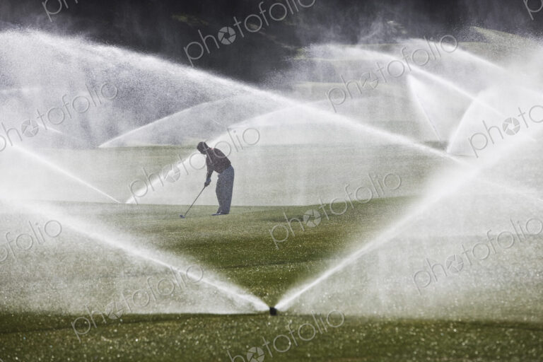 A golfer hitting his second shot on the fairway when the sprinkler system turns on.