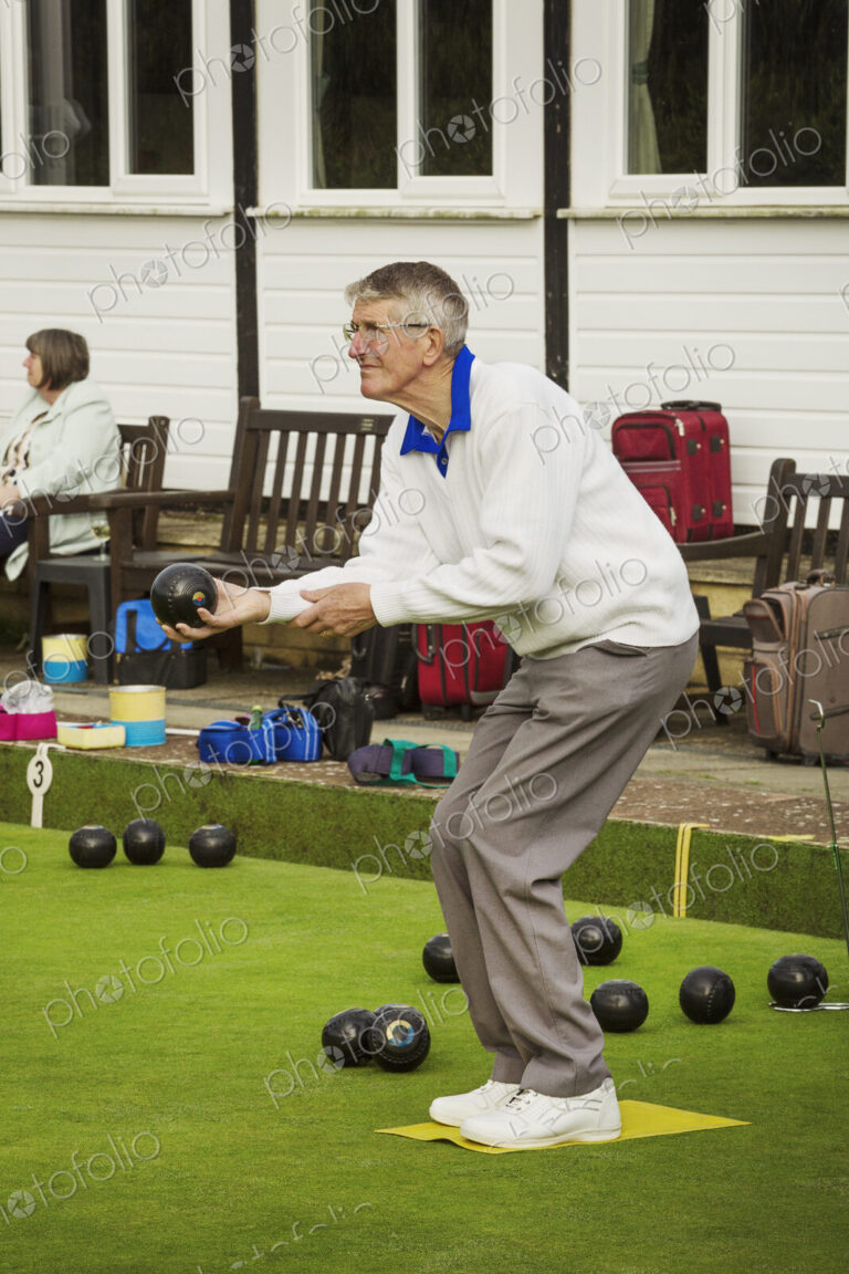 A grey haired man standing on a yellow mat on a lawn bowls green, bending his knees…