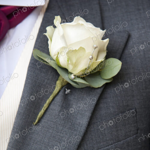 A groom in a grey jacket and white shirt, with a white rose buttoniere, in his buttonhole.