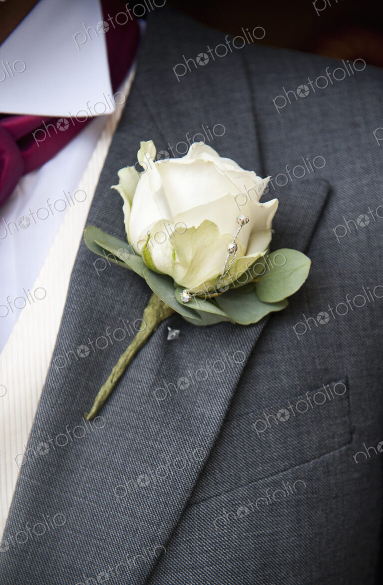 A groom in a grey jacket and white shirt, with a white rose buttoniere, in his buttonhole.