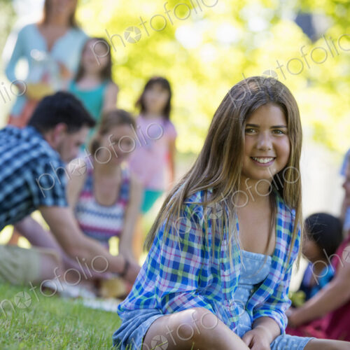 A group of adults and children sitting on the grass under the shade of a tree. A…