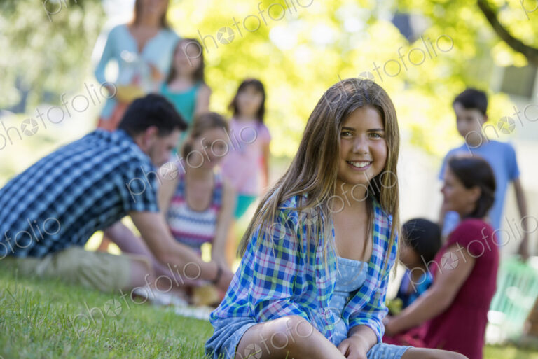 A group of adults and children sitting on the grass under the shade of a tree. A…