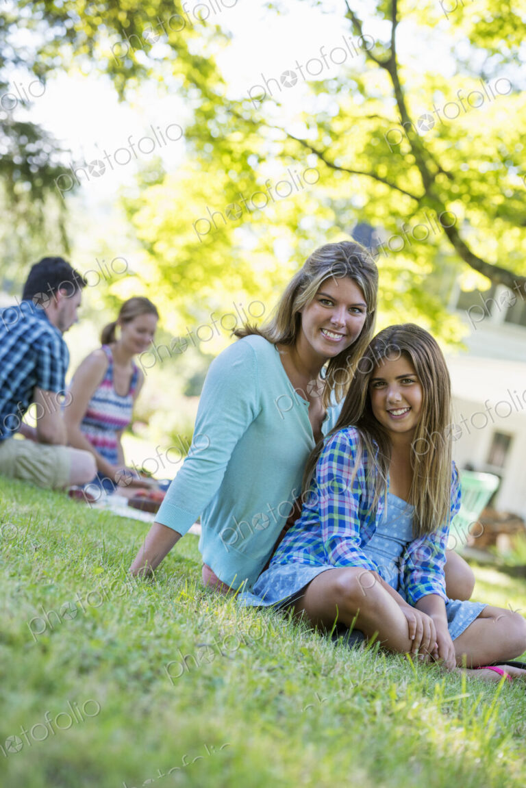 A group of adults and children sitting on the grass under the shade of a tree. A…