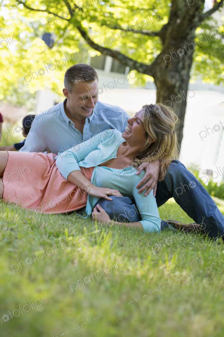A group of adults and children sitting on the grass under the shade of a tree. A…
