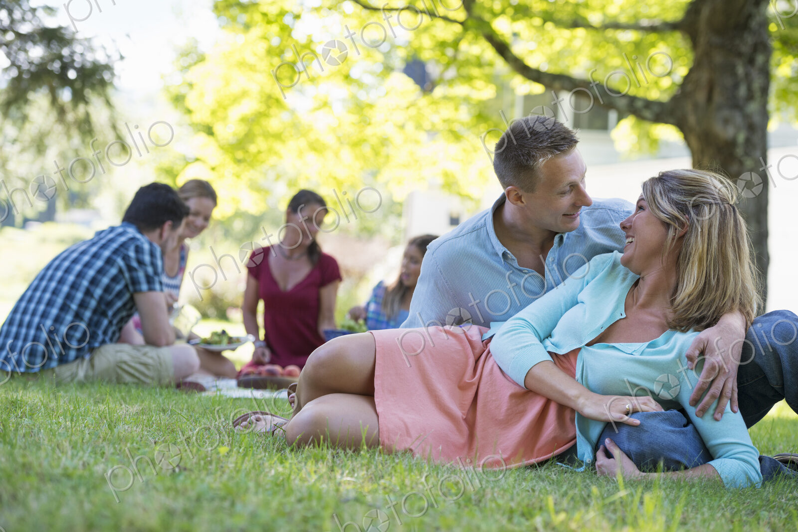A group of adults and children sitting on the grass under the shade of a tree. A…