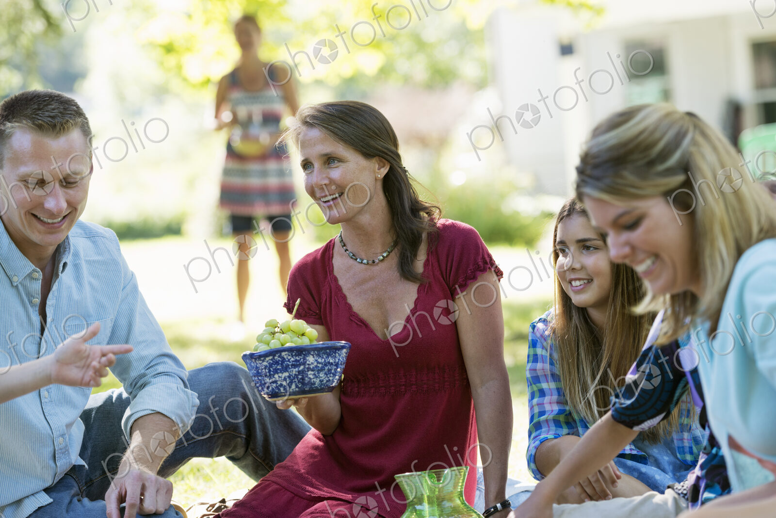 A group of adults and children sitting on the grass under the shade of a tree. A…