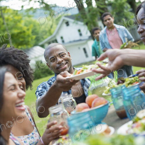 A group of adults and young people at a meal in the garden of a farmhouse. Passing…