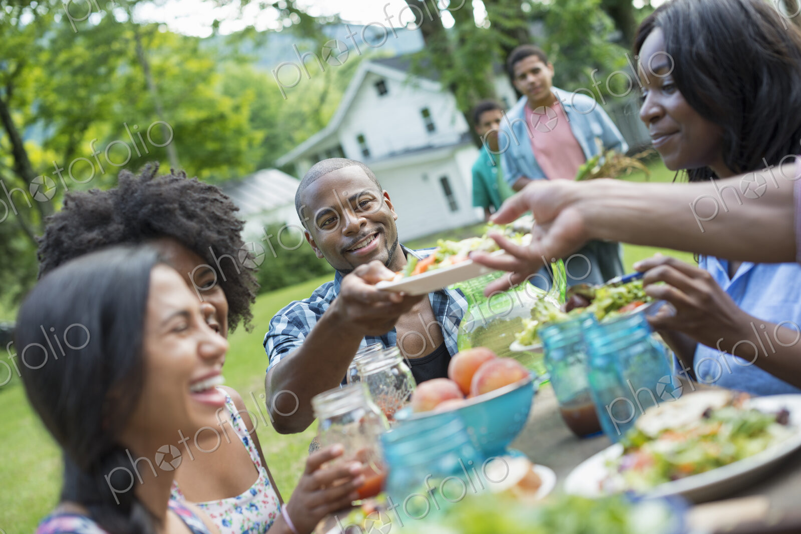 A group of adults and young people at a meal in the garden of a farmhouse. Passing…
