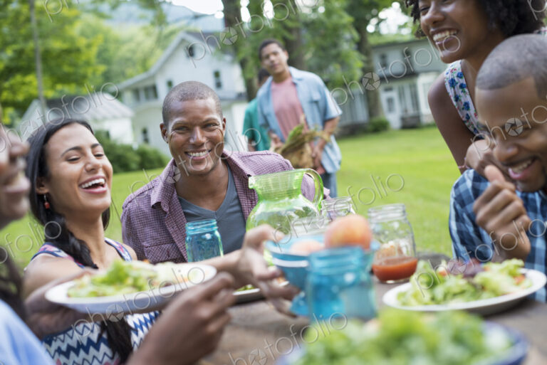 A group of adults and young people at a meal in the garden of a farmhouse. Passing…