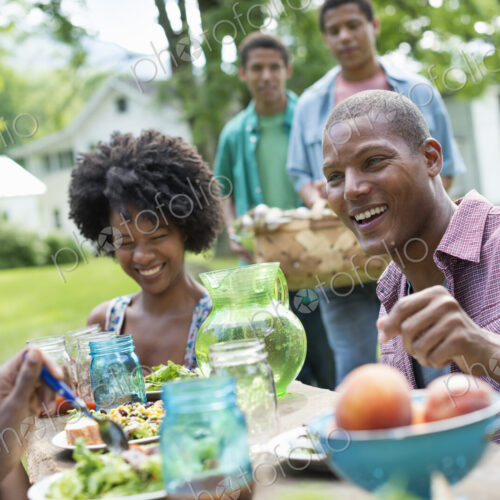 A group of adults and young people at a meal in the garden of a farmhouse. Passing…