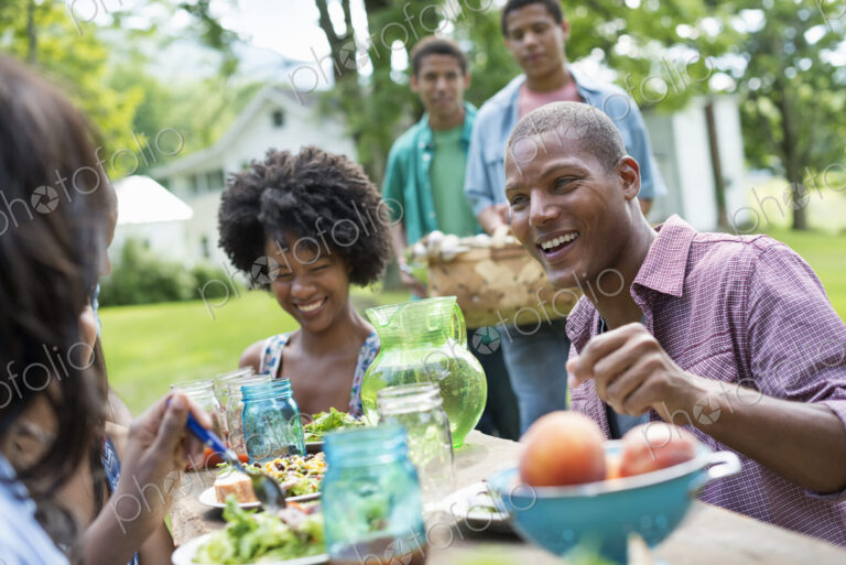 A group of adults and young people at a meal in the garden of a farmhouse. Passing…