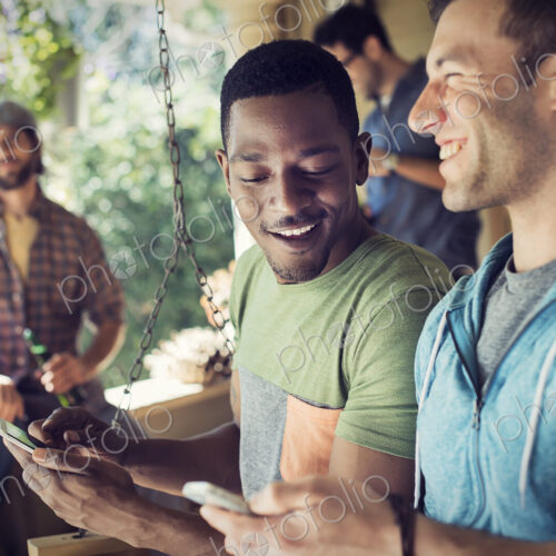 A group of friends, men and women at a house party, two men looking at their smart phones.