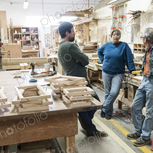 A group of mixed race carpenters discussing a project at a work station in a large… A group of mixed race carpenters discussing a project at a work station in a large…