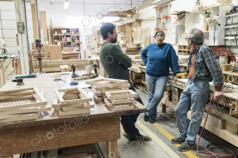A group of mixed race carpenters discussing a project at a work station in a large…