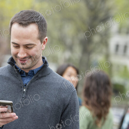 A group of people in a city park. A man in a grey sweater, checking his phone.