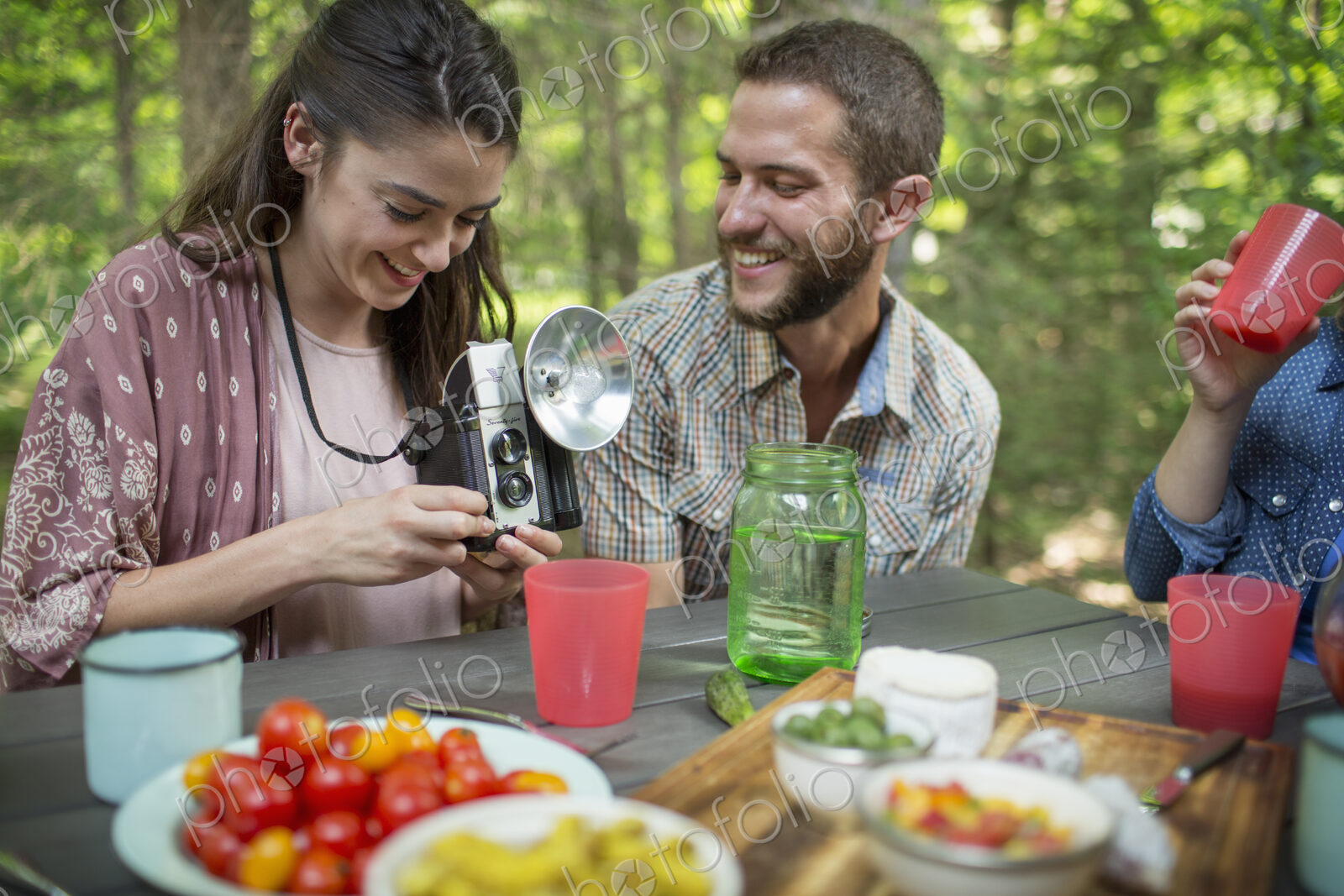 A group of people seated at a meal around a table outdoors. A couple side by side, one…
