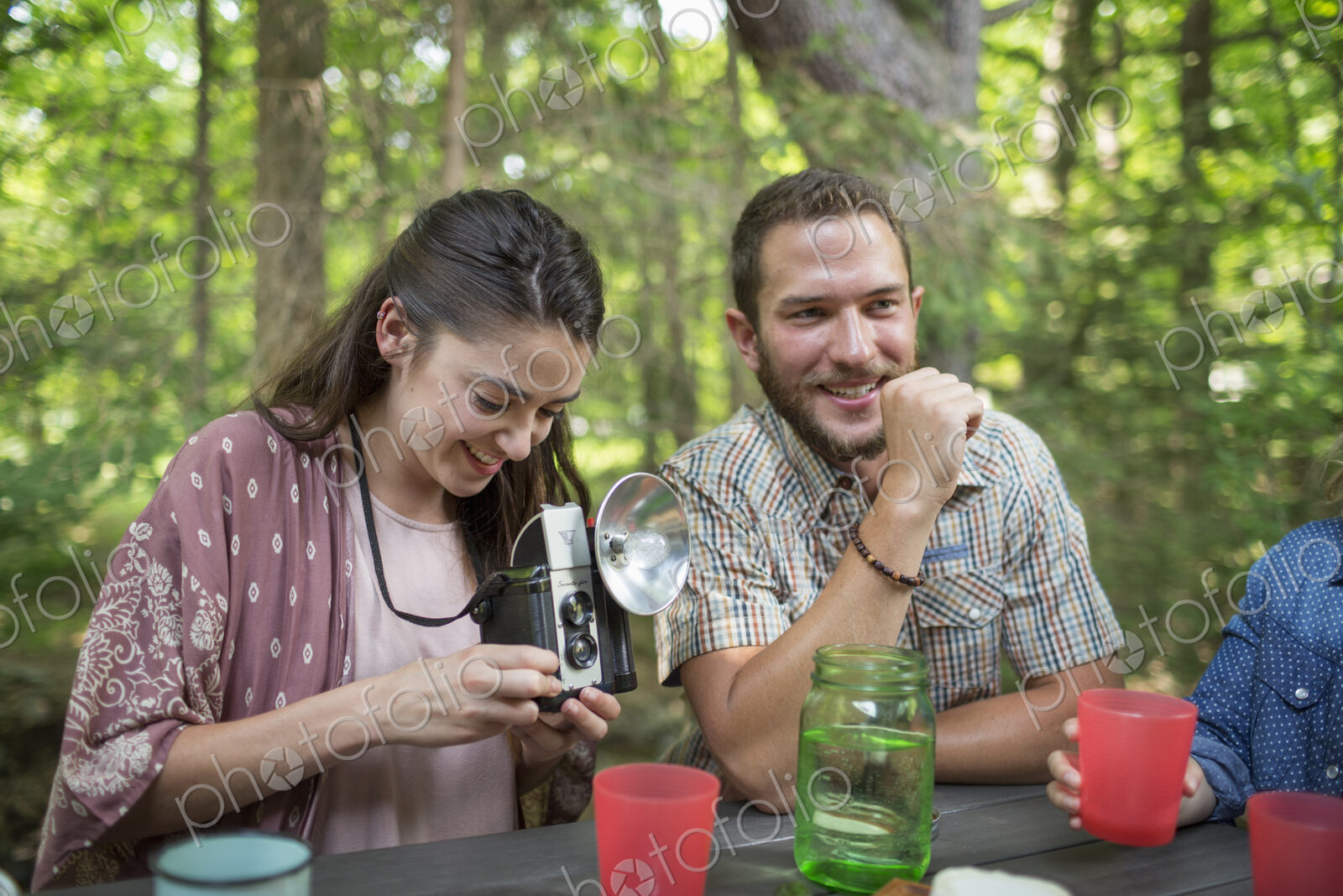 A group of people seated at a meal around a table outdoors. A couple side by side, one…