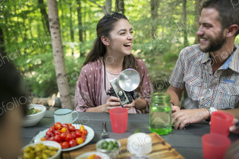 A group of people seated at a meal around a table outdoors. A couple side by side, one…