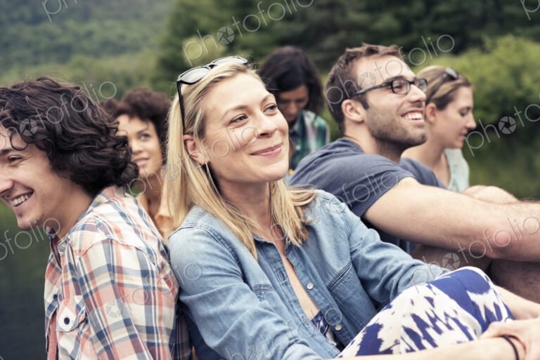 A group of women and men seated on a jetty by a lake in the countryside.