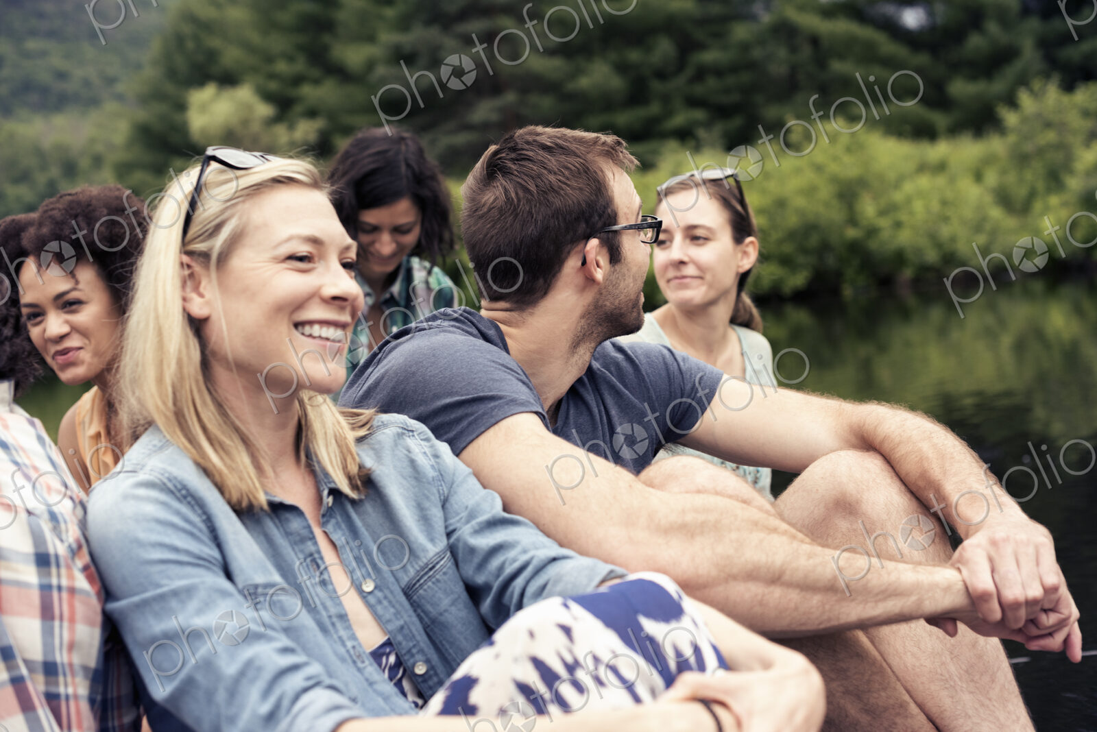 A group of women and men seated on a jetty by a lake in the countryside.