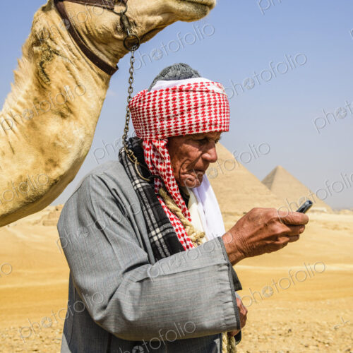 A guide in traditional Arab dress, robe and turban using a mobile phone, and his camel.