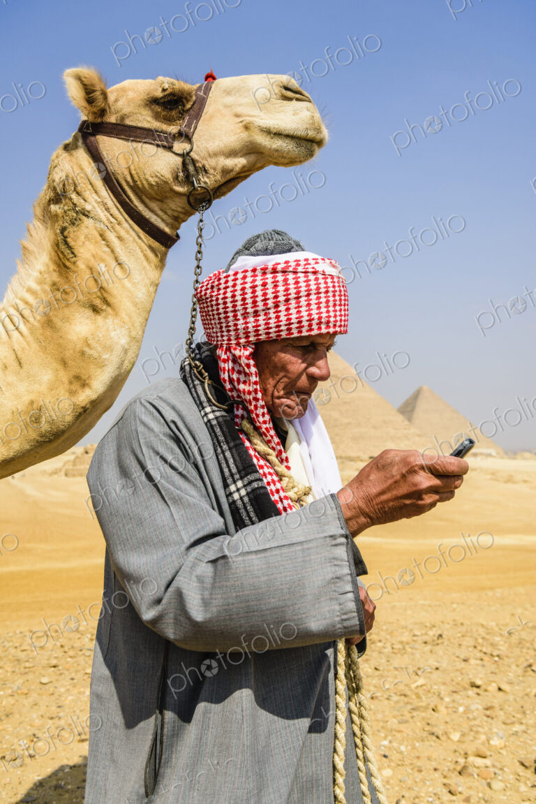 A guide in traditional Arab dress, robe and turban using a mobile phone, and his camel.