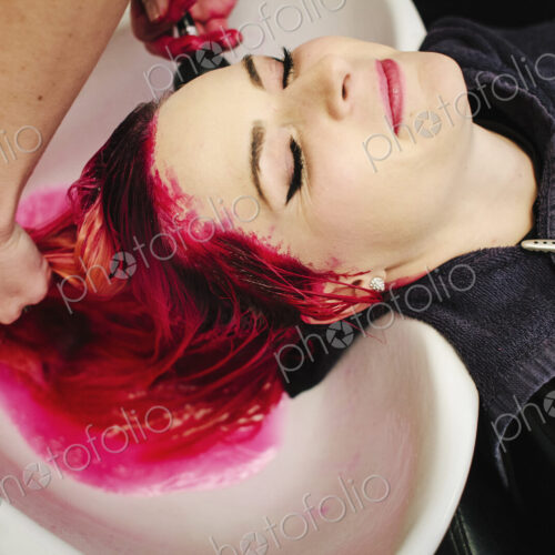 A hair salon client having red hair dye rinsed from her hair over a basin.