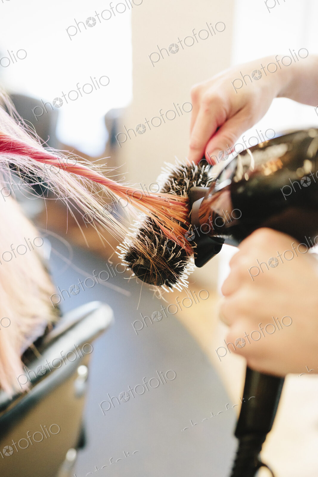 A hair stylist blowdrying a client’s long straight pink hair using a round bristle brush.