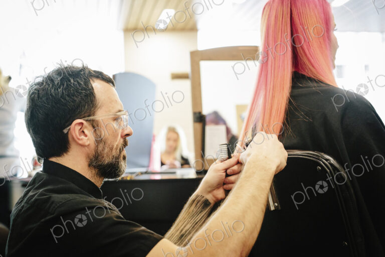 A hair stylist with a client, trimming the ends of her long dyed pink straight hair.