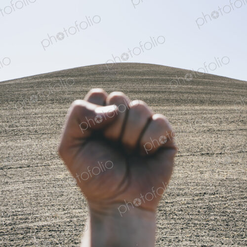 A hand bunched up and making fist, making a gesture. A ploughed field and farmland in…
