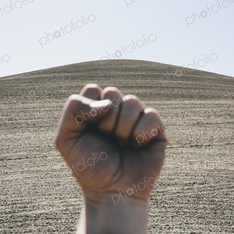 A hand bunched up and making fist, making a gesture. A ploughed field and farmland in…