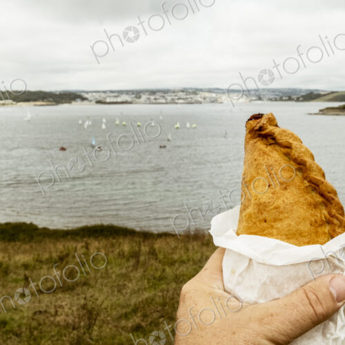 A hand holding a Cornish pasty baked pastry with a crimped edge, seated overlooking a…