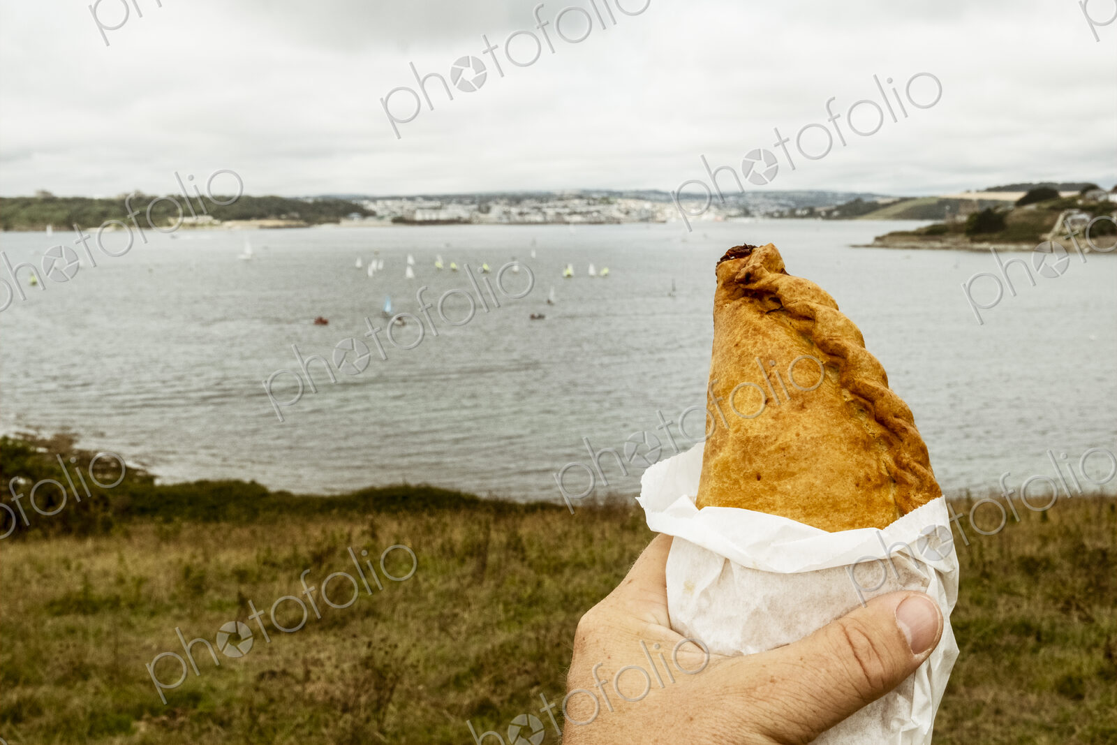 A hand holding a Cornish pasty baked pastry with a crimped edge, seated overlooking a…