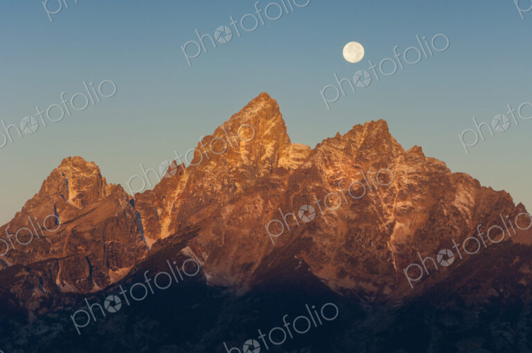 A jagged mountain range in the Grand Teton national park at night, with a full moon in…