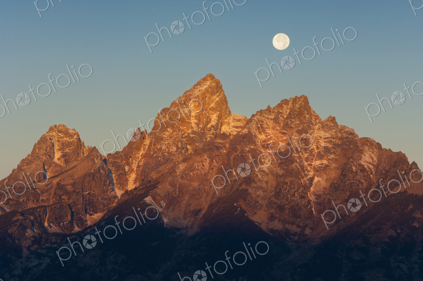 A jagged mountain range in the Grand Teton national park at night, with a full moon in…
