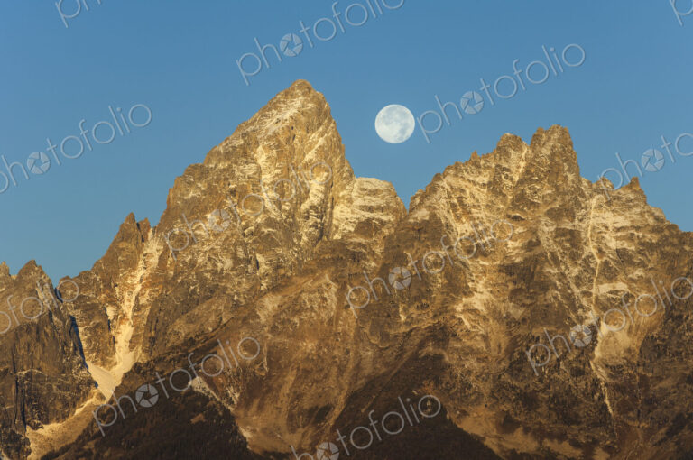 A jagged mountain range in the Grand Teton national park at night, with a full moon in…