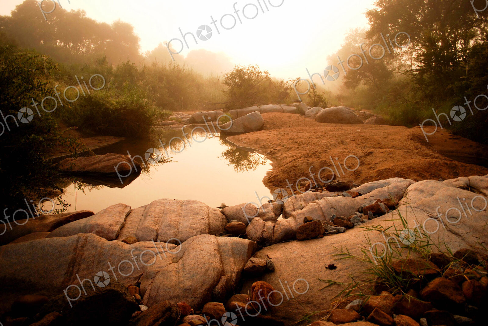 A landscape, a waterhole surrounded by boulders and sand at dusk, with mist in the…