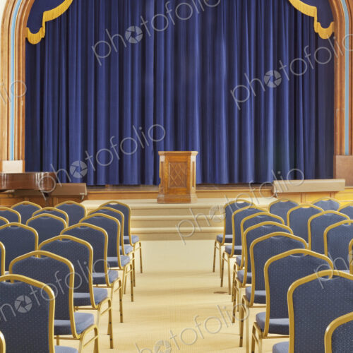 A large auditorium in a historic building, with blue chairs. View to the stage and podium.