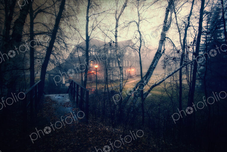 A large house at dusk with lights glowing. Woodland. A footbridge.
