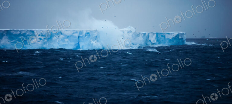 A large iceberg with steep sides, floating on the water. Waves and spray rising.…
