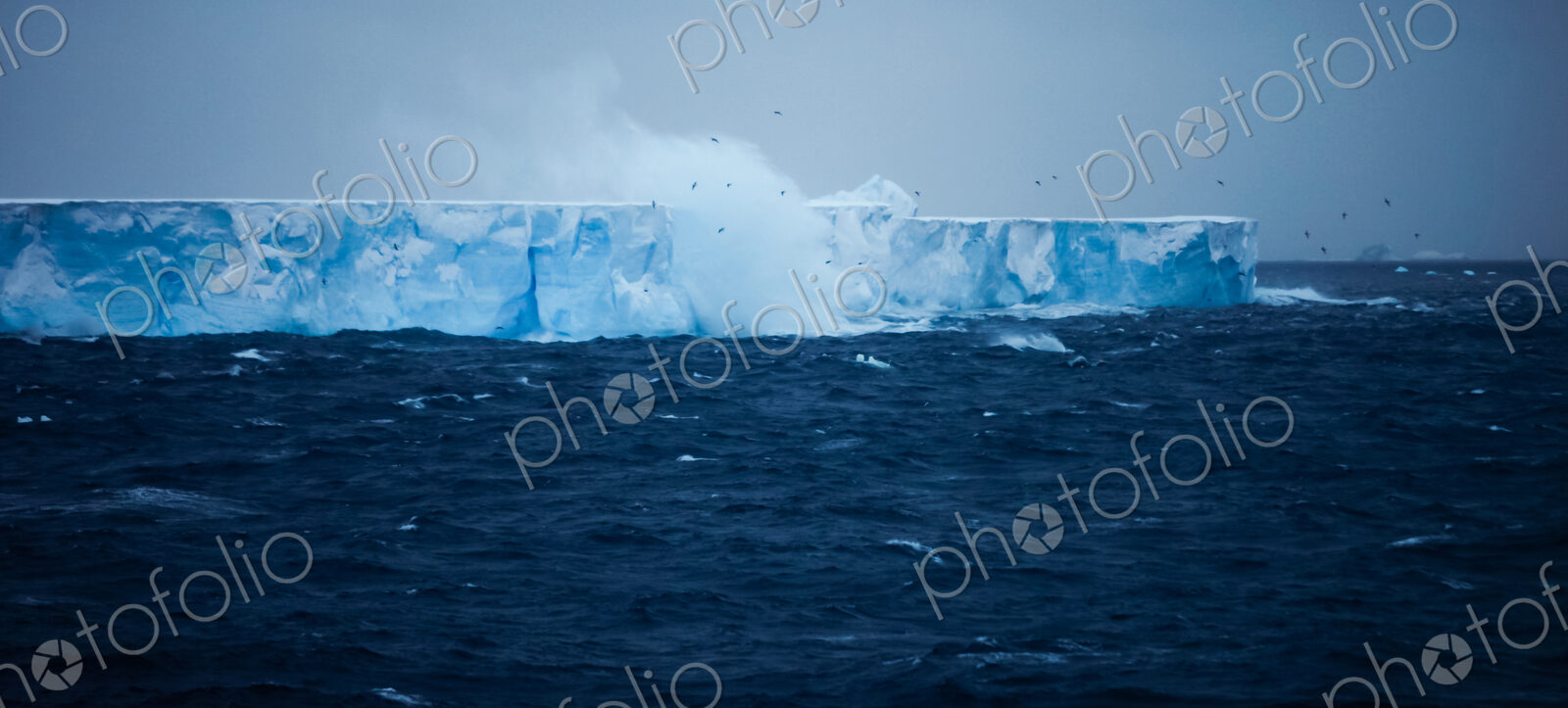 A large iceberg with steep sides, floating on the water. Waves and spray rising.…