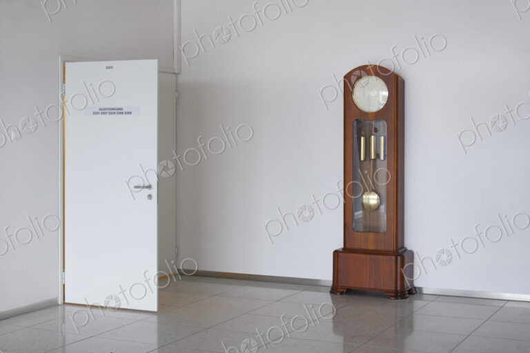 A large modern grandfather clock with weights and pendulum in a white empty room.
