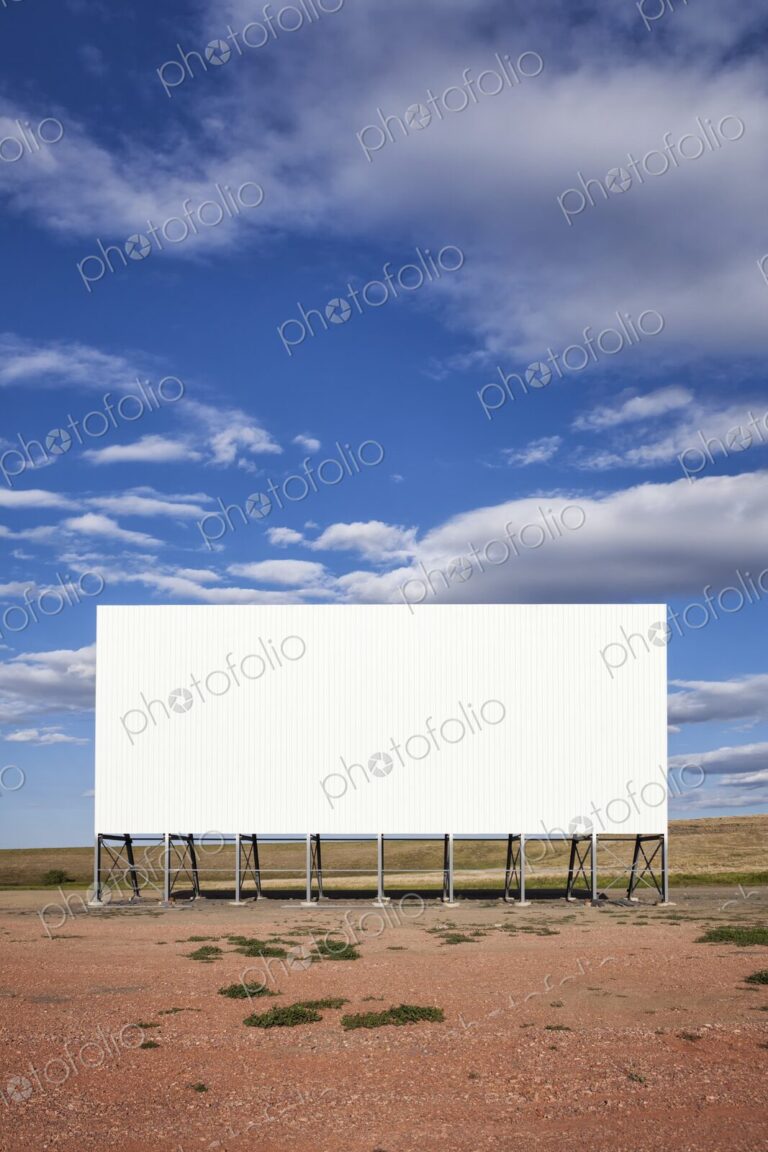 A large white drive in movie screen stands on the prairie beneath a wide blue sky with…