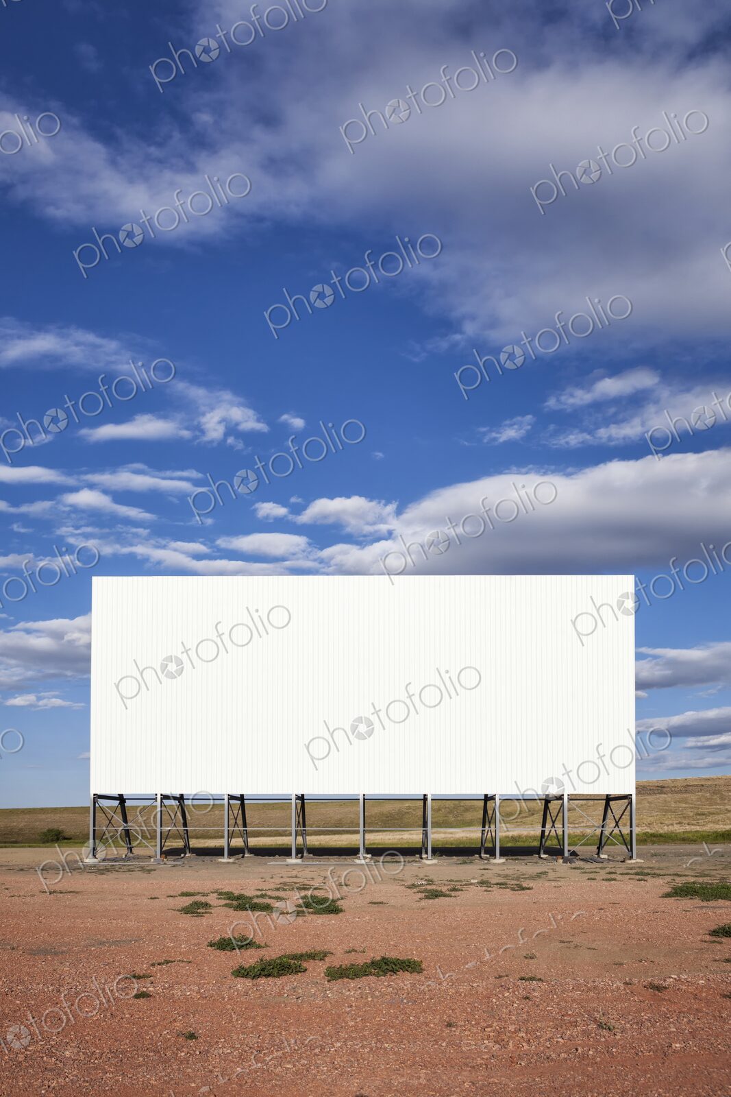 A large white drive in movie screen stands on the prairie beneath a wide blue sky with…