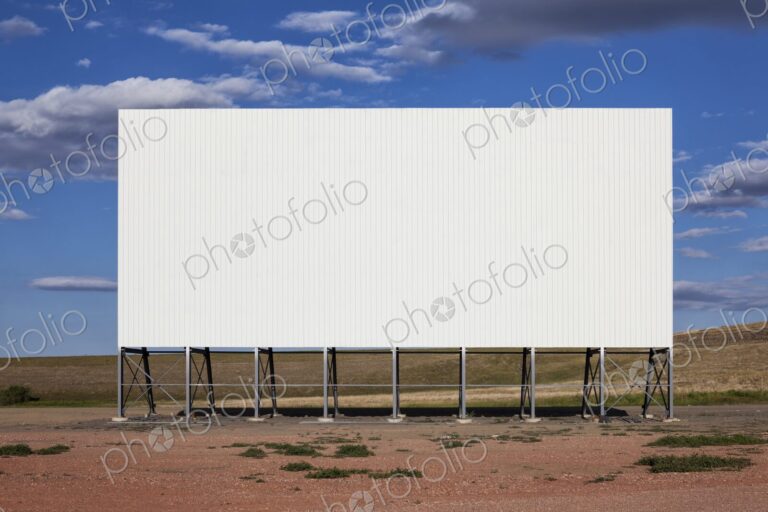 A large white drive in movie screen stands on the prairie beneath a wide blue sky with…